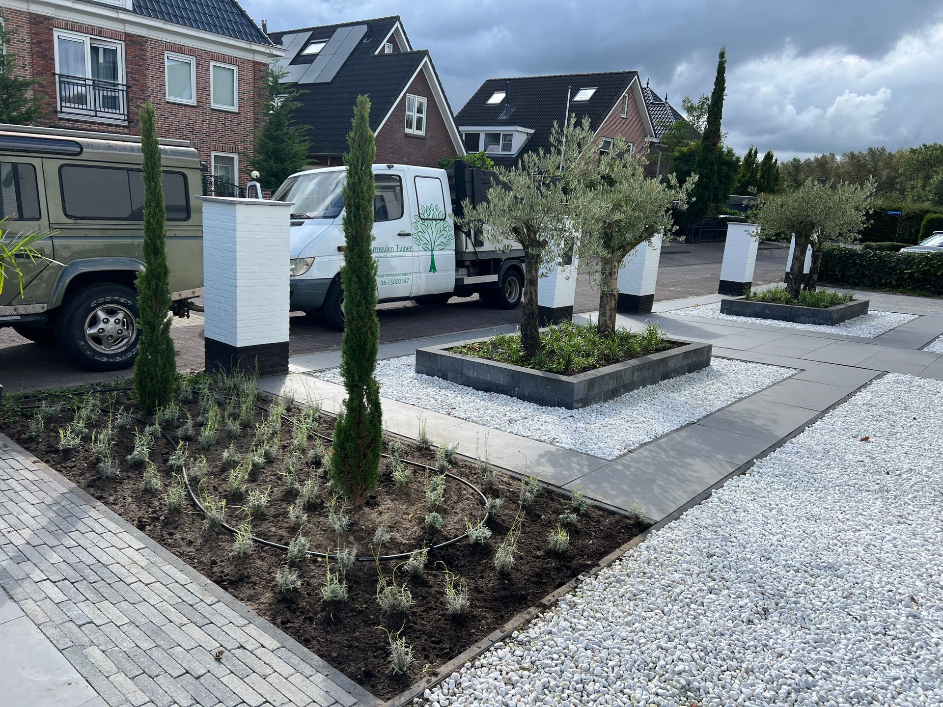 Een wit busje staat geparkeerd op een oprit naast een tuin.