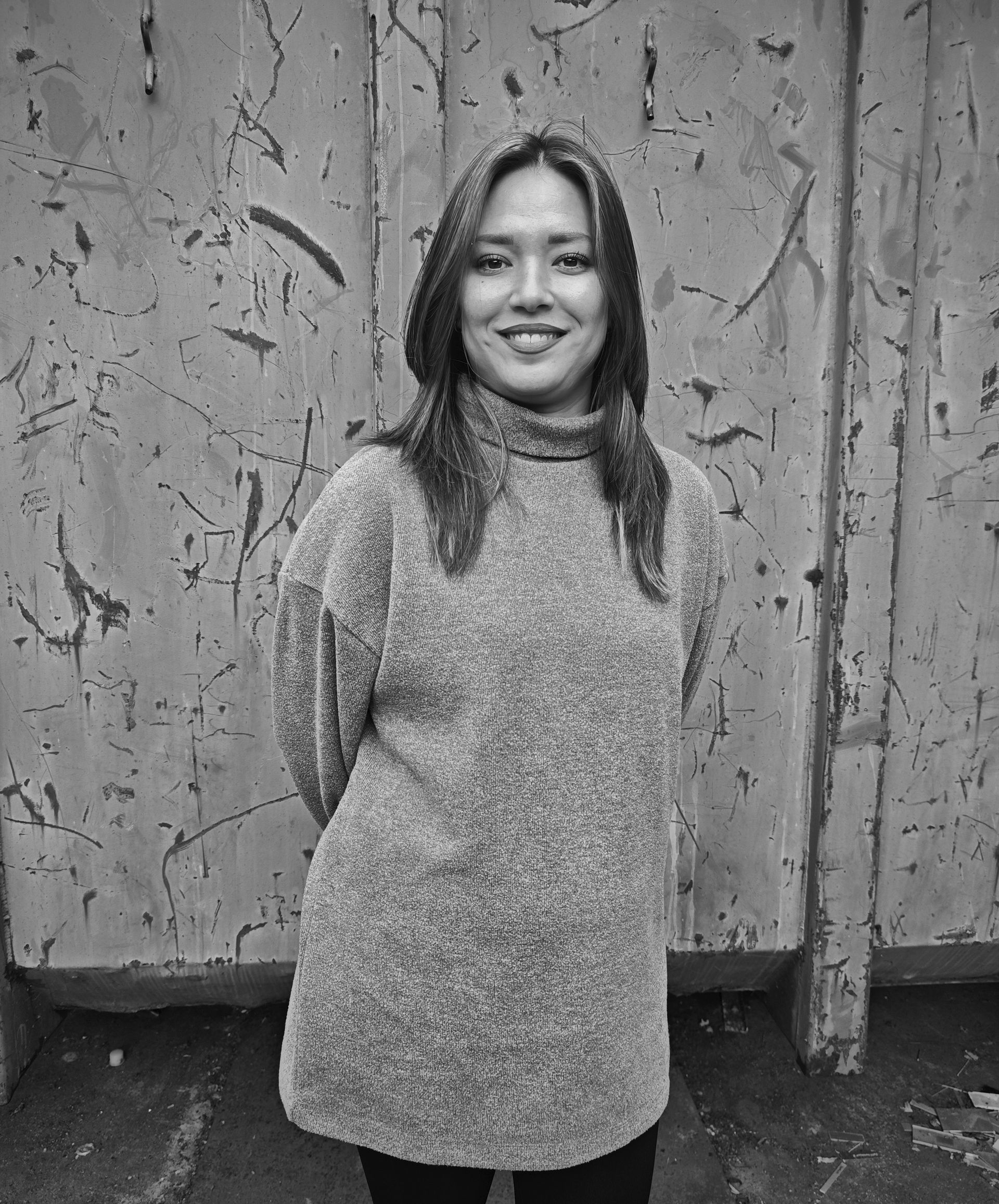 Eine lächelnde Frau steht vor einer strukturierten Metallwand und trägt einen Pullover und eine dunkle Hose.