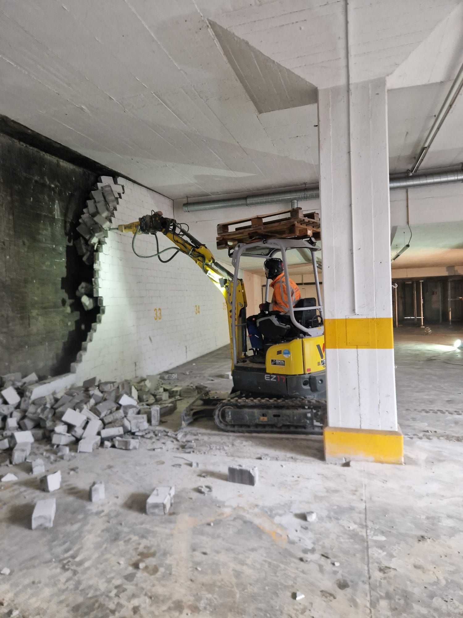 Ein kleiner gelber Bagger reißt eine Betonwand in einem Parkhaus ein. Trümmer liegen auf dem Boden.