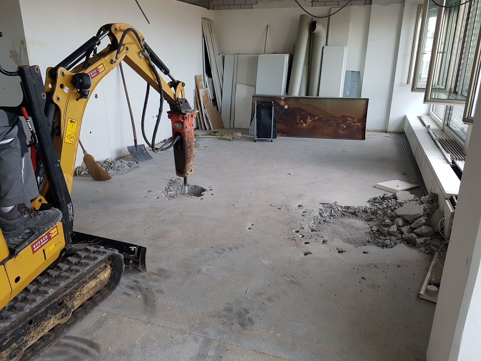 Ein kleiner Bagger arbeitet an einem Betonboden in einem Raum.