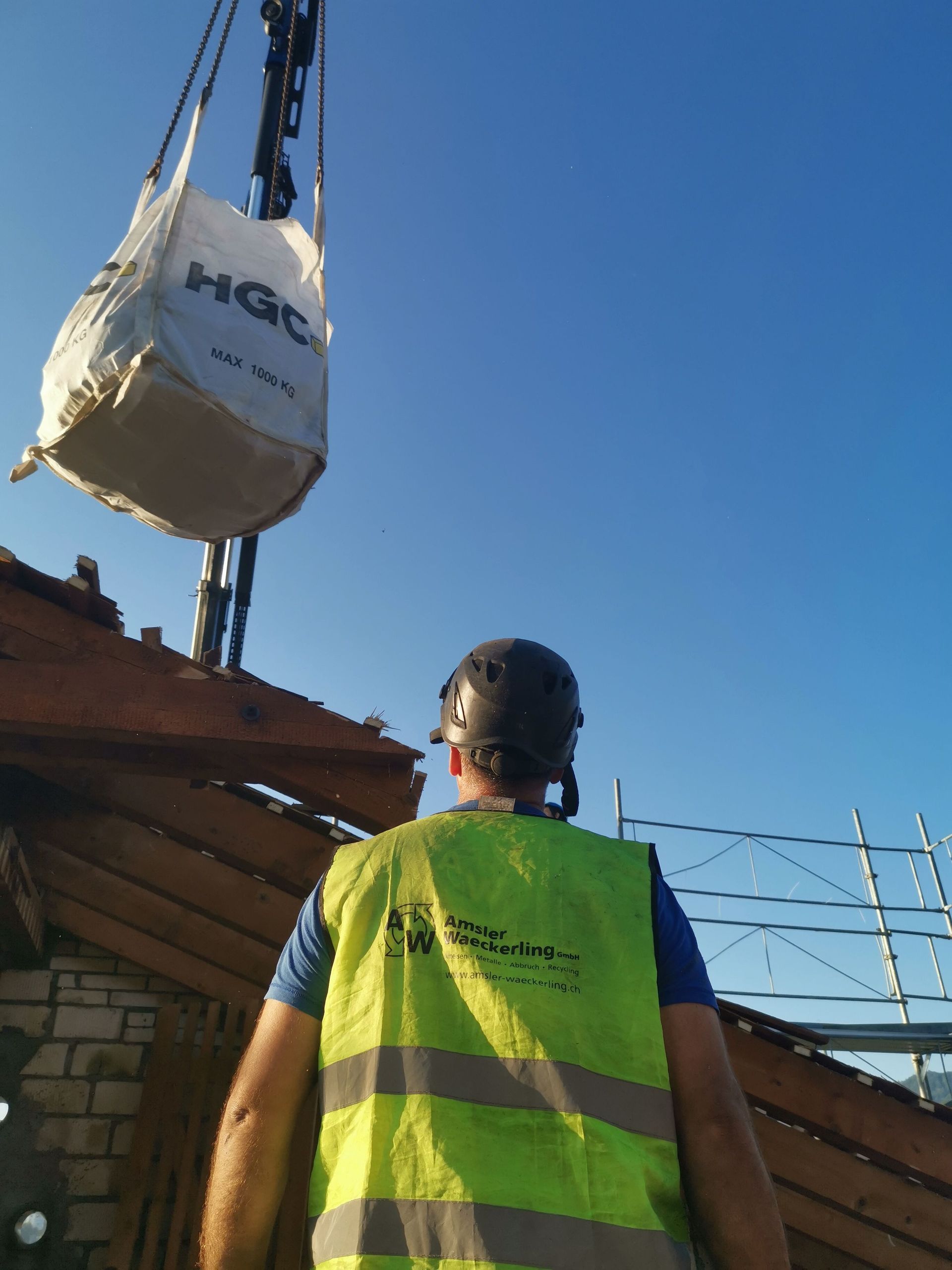 Ein Mann in einer gelben Weste steht vor einem Kran und hebt einen Sack hoch.