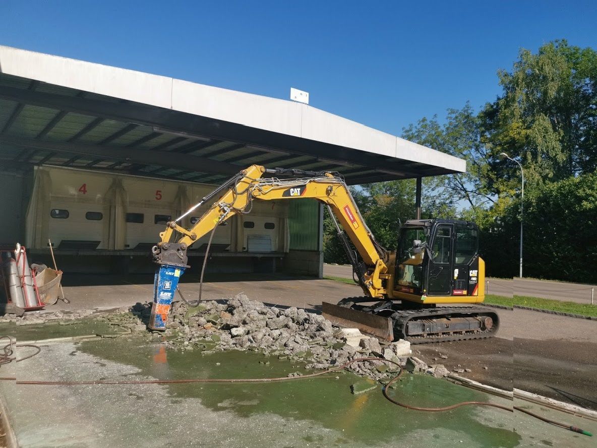 Ein gelber Bagger bricht Beton vor einem Gebäude auf.