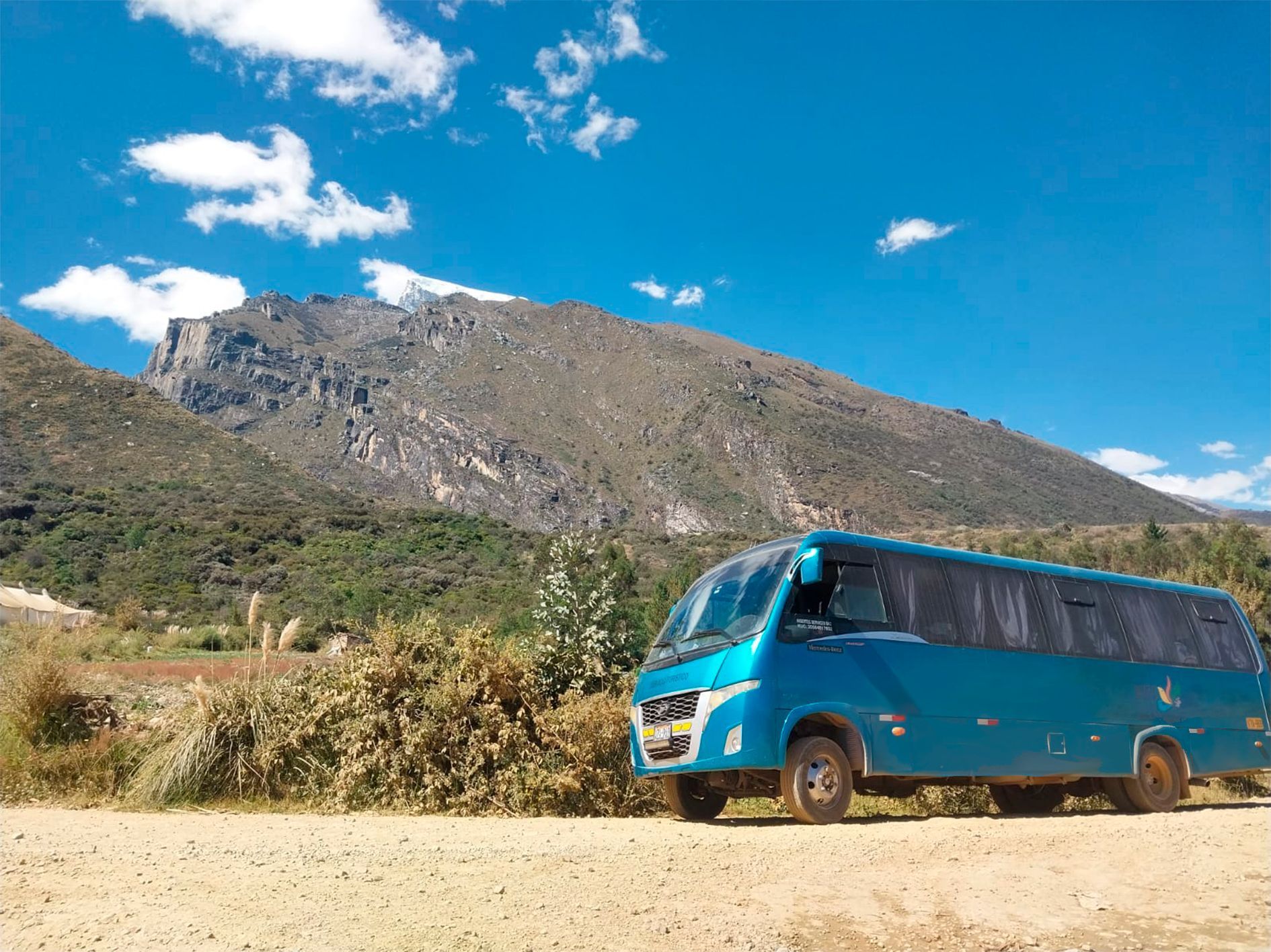 Un autobús azul está estacionado al costado de un camino de tierra frente a una montaña.