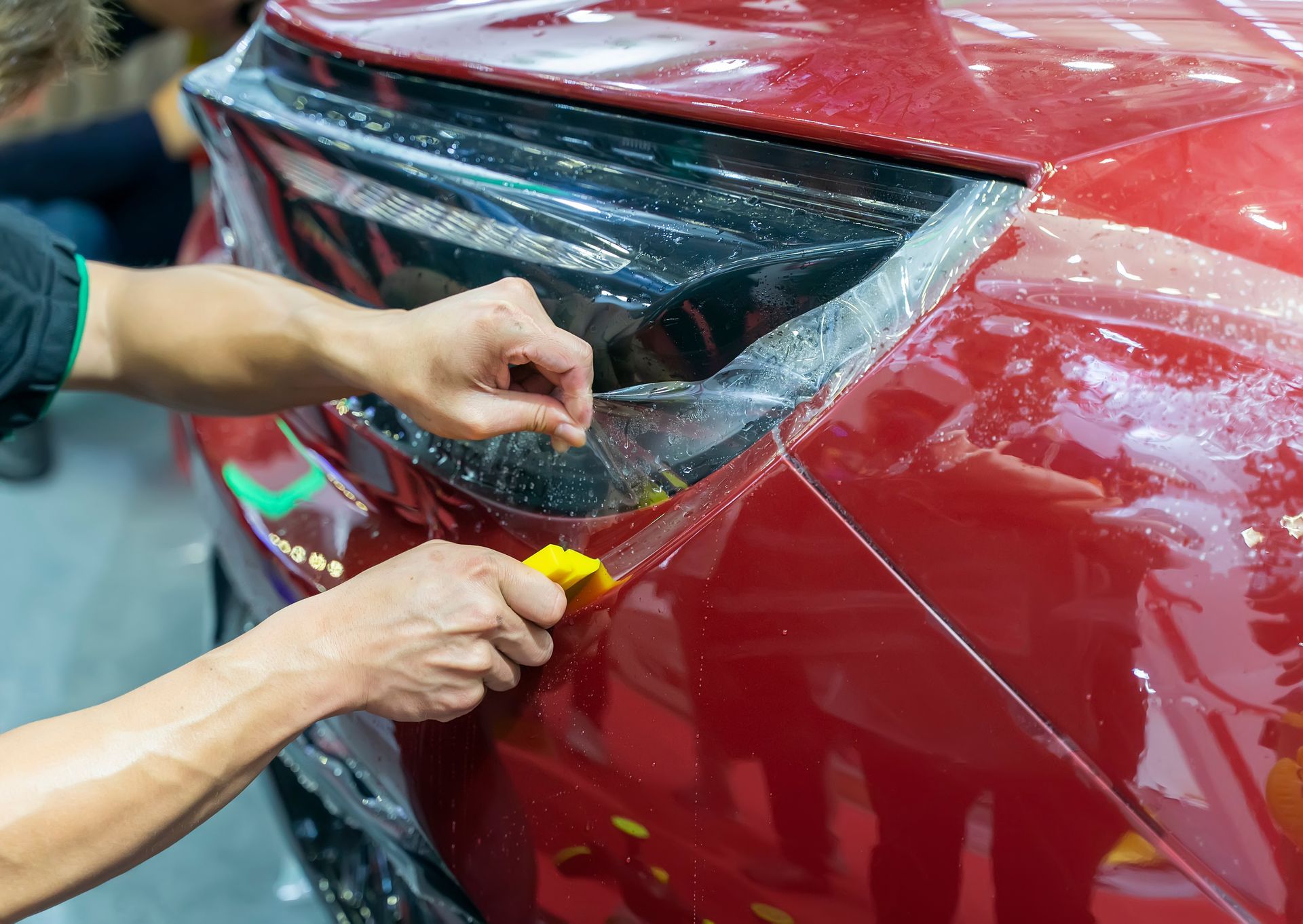 A man is applying a protective film to the headlights of a red car.