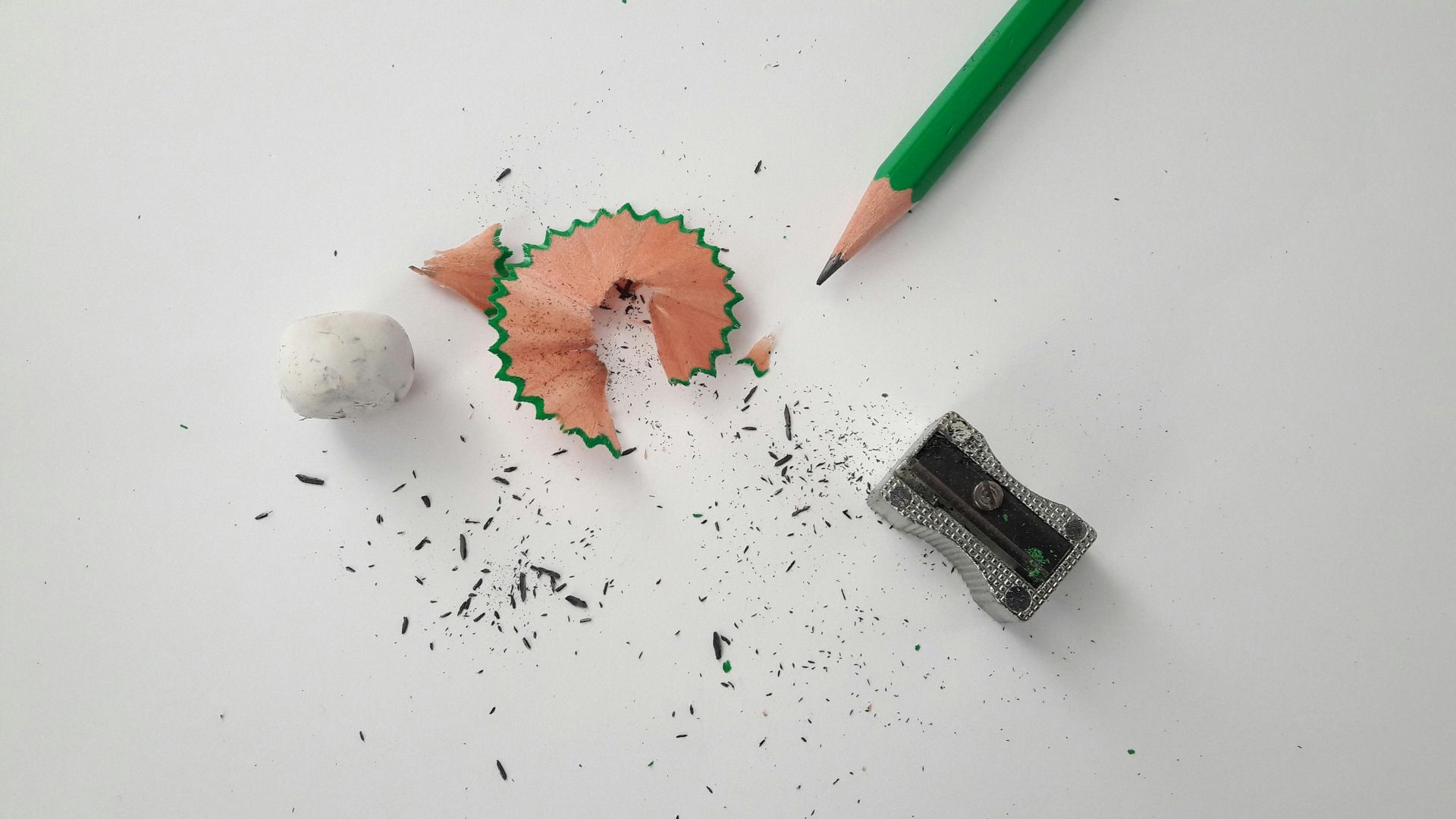 A green pencil, a metal sharpener, a wood shaving, and a small eraser on a plain white surface.