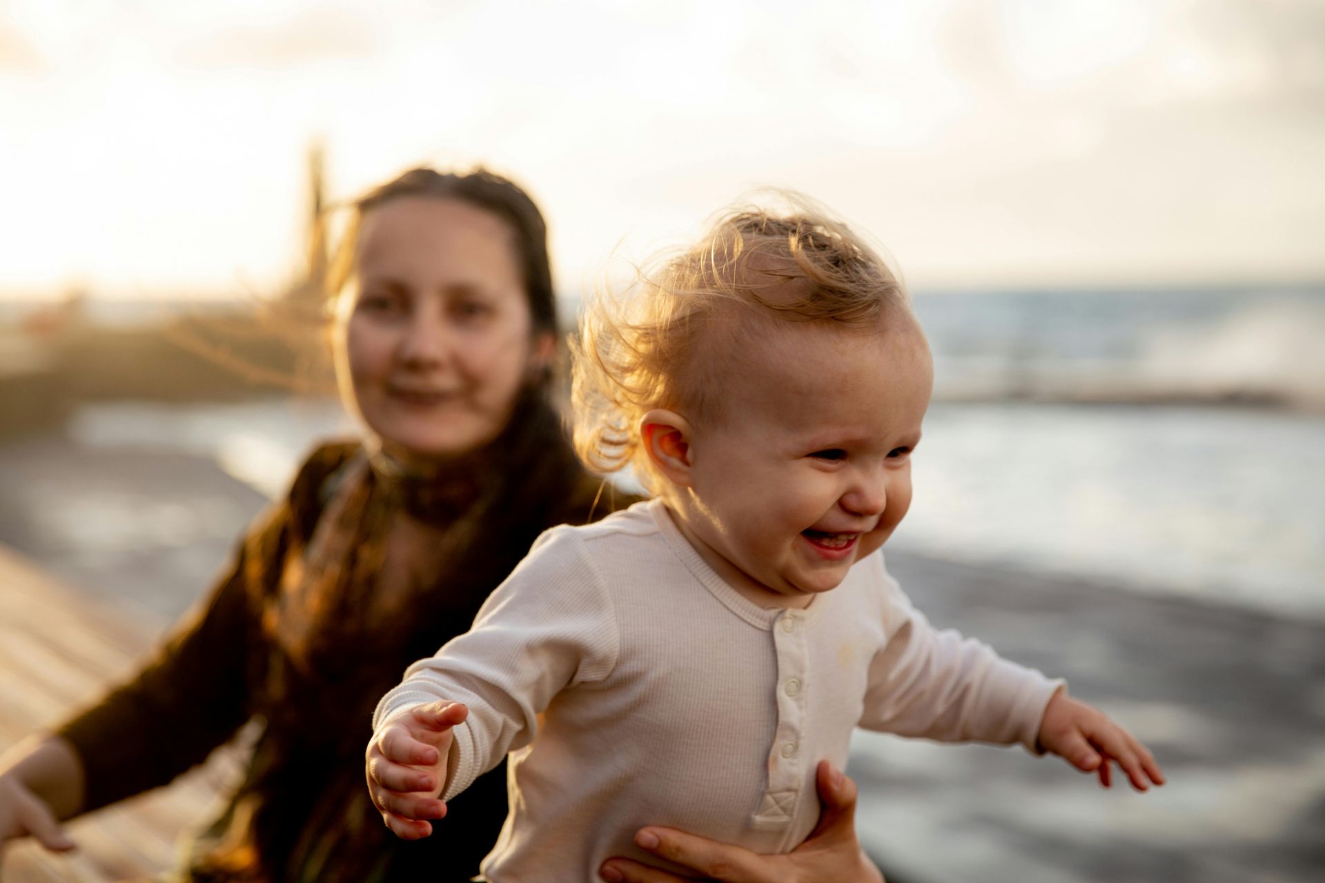 A person smiles while holding a laughing infant on a beach during a golden sunset.