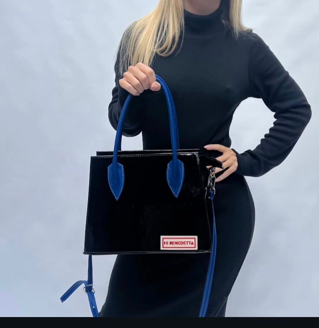 Una mujer con un vestido negro sostiene un bolso negro con asas azules.