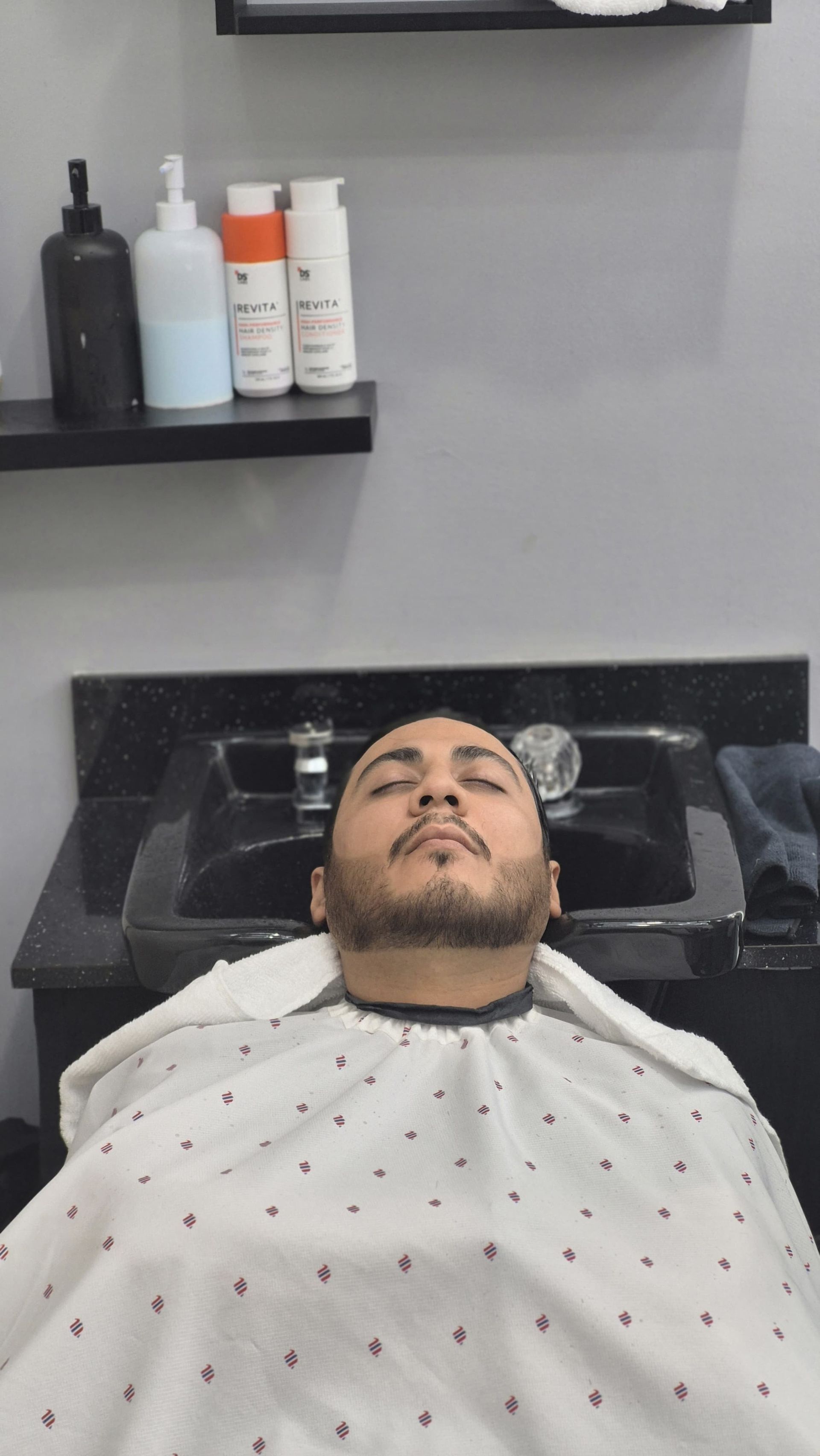 Man in barber chair, face up, draped in a barber cape. Sink and products are in background.