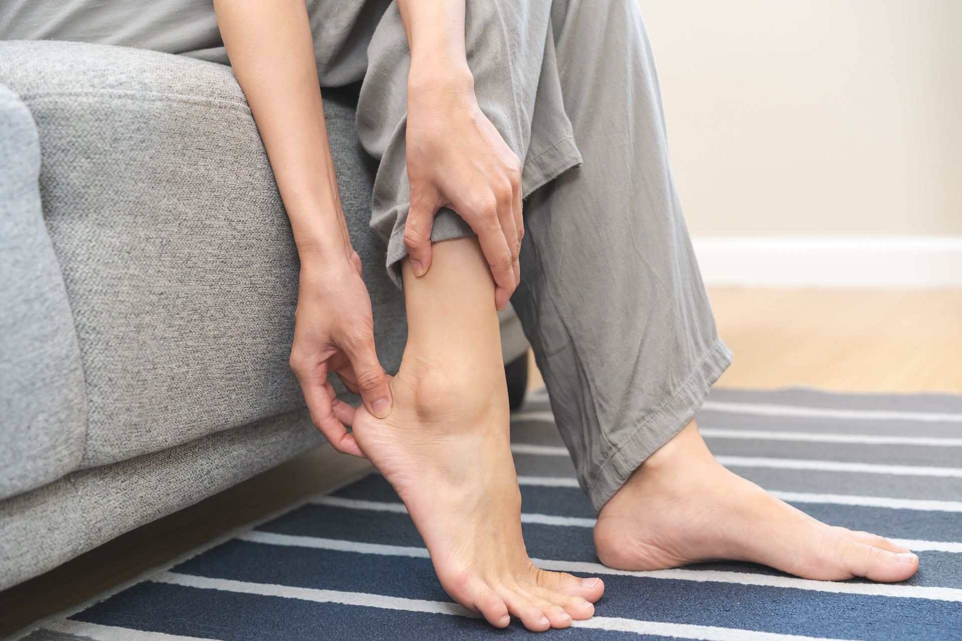Person seated, holding their ankle, possibly in pain. Gray pants, striped rug, light background.