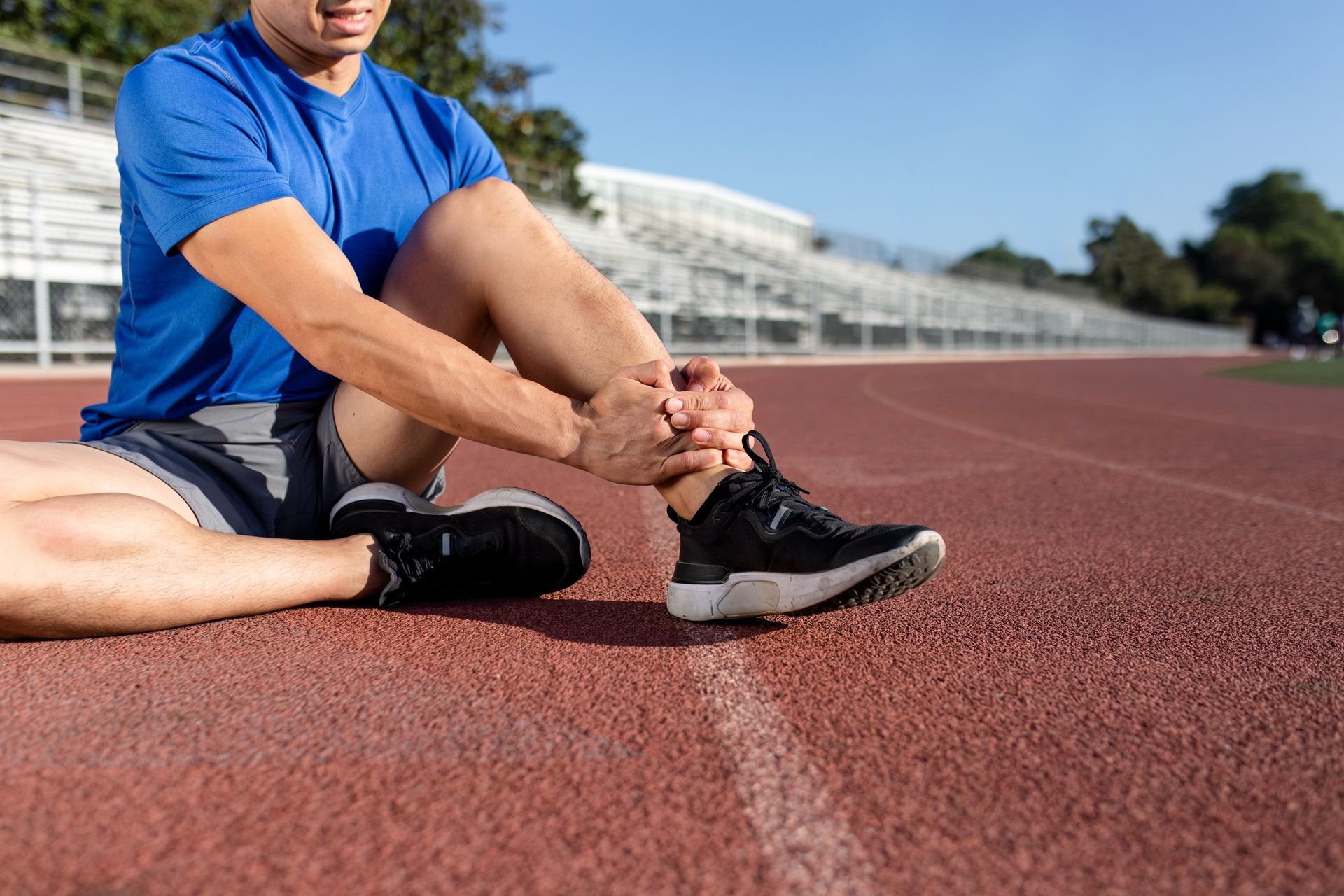 Athlete on a track holding his ankle, possibly injured, wearing black athletic shoes and a blue shirt.