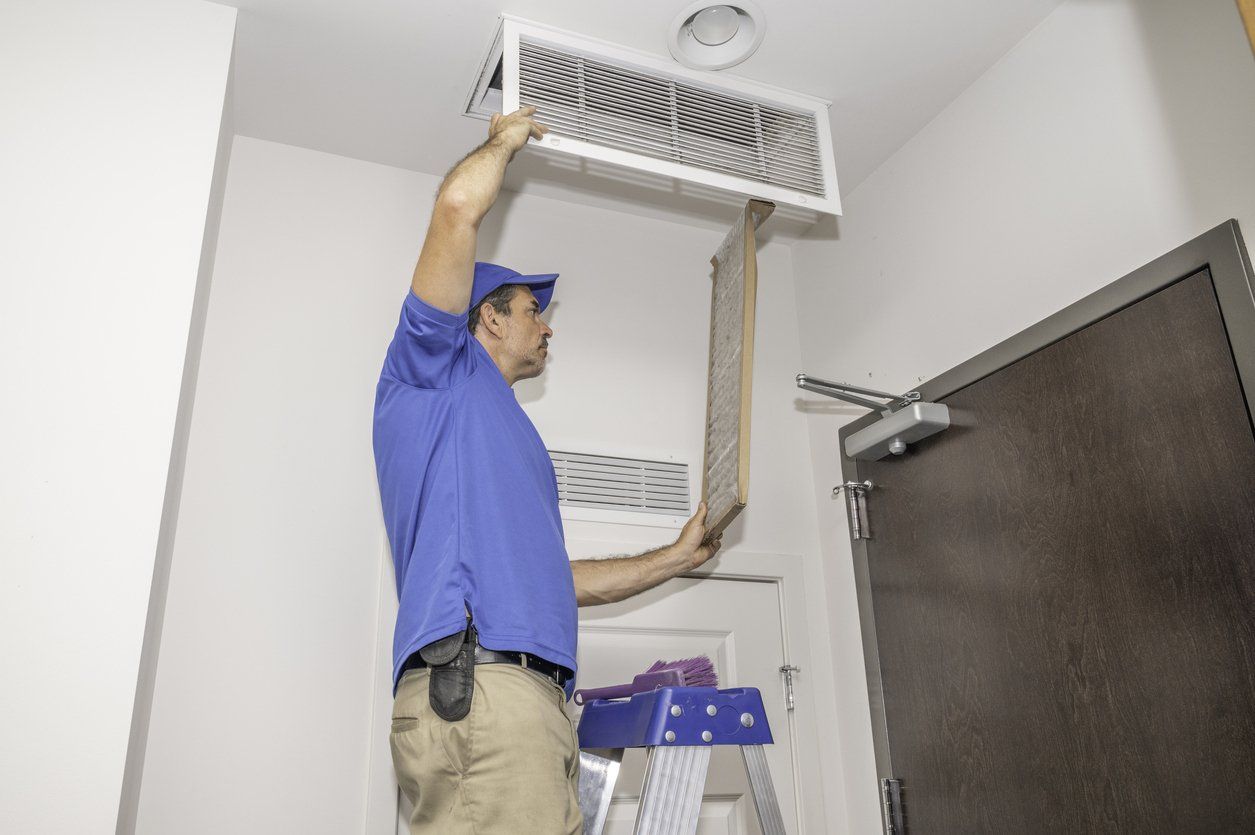 A male maintenance technician replacing a dirty air filter while standing on a ladder.