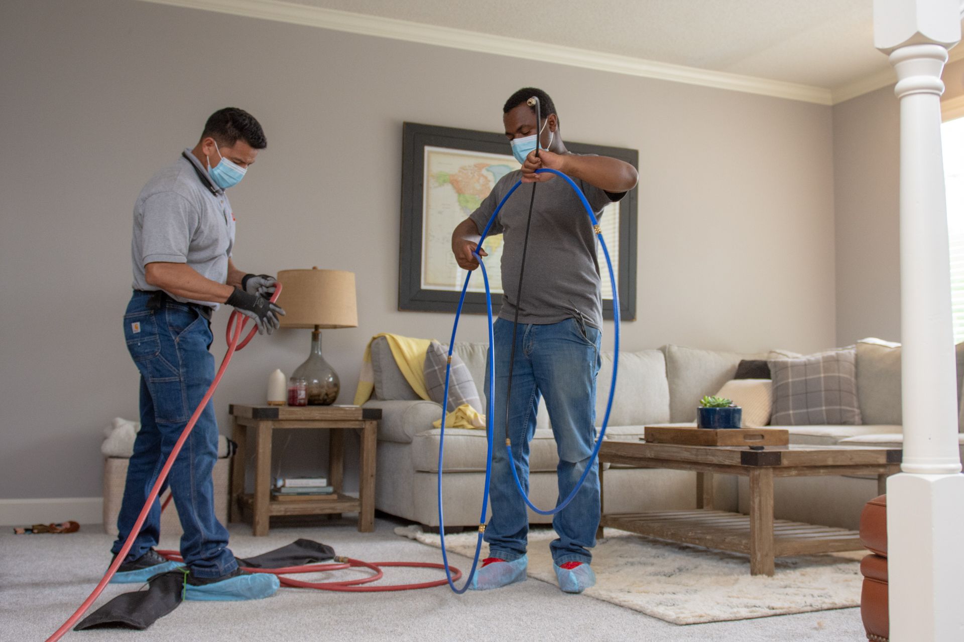 Two men wearing masks are working on a carpet in a living room.