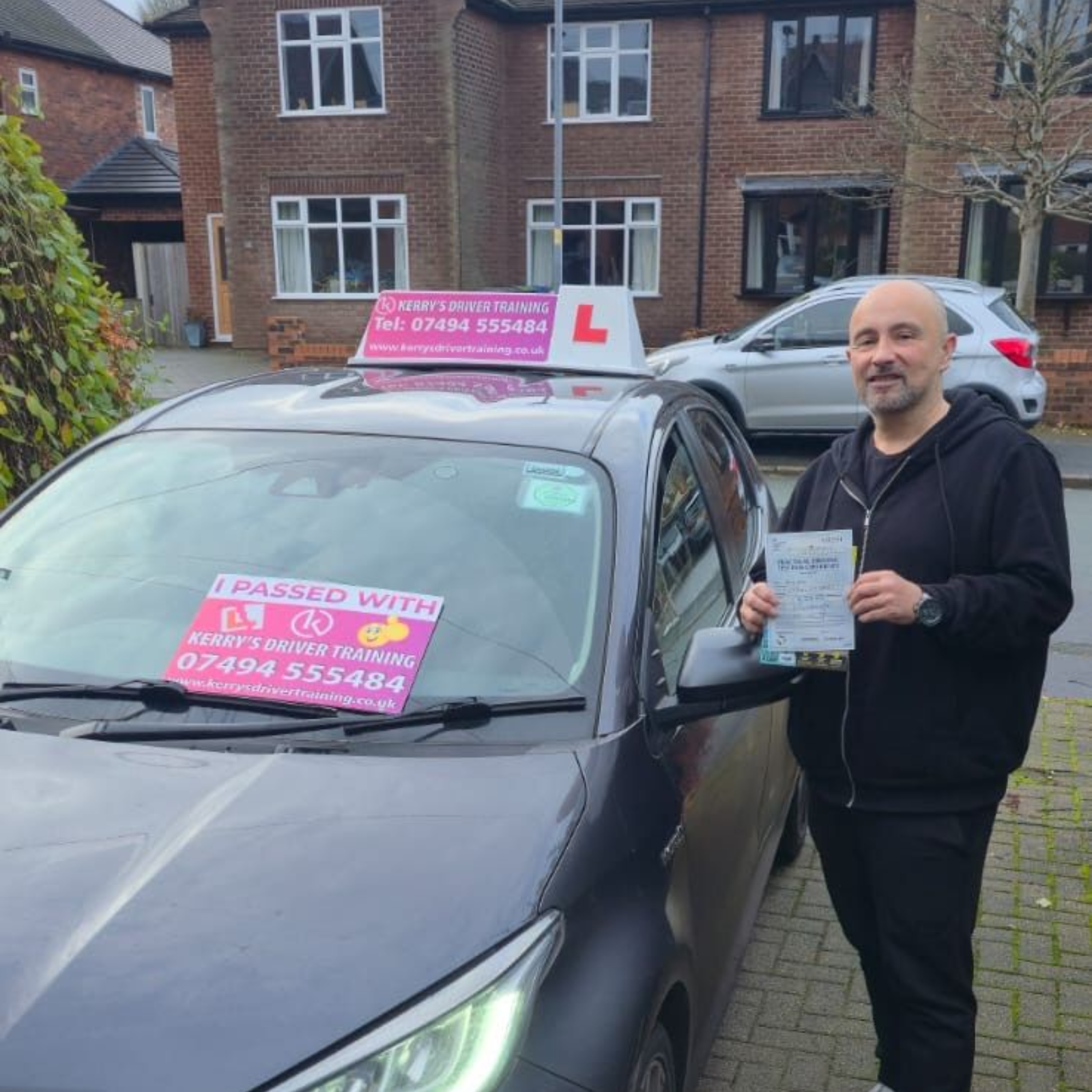 Man holding a driving license next to a dark gray car with pink L plates in front of a house.