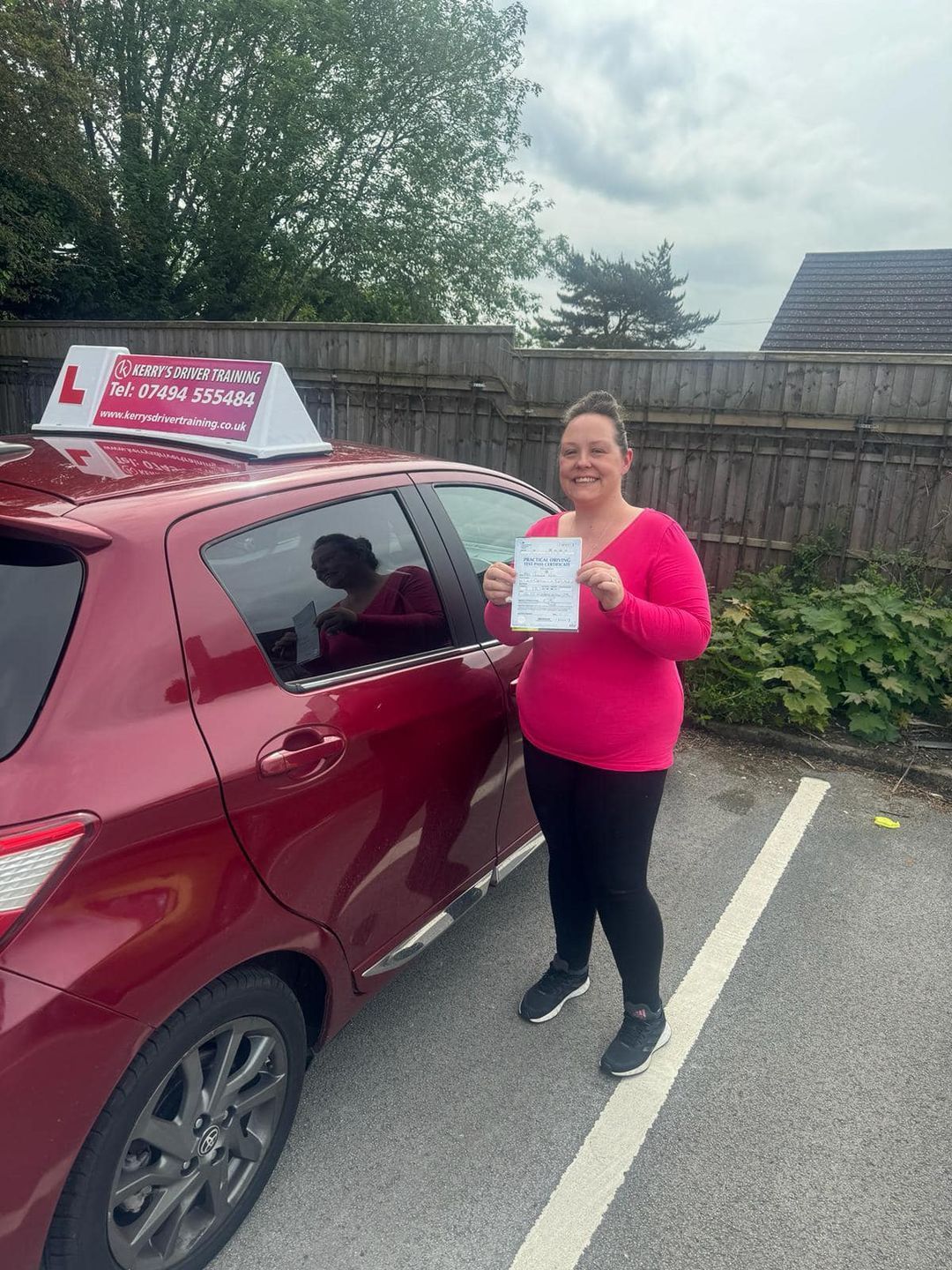 A woman is standing in front of a red car holding a driving certificate.