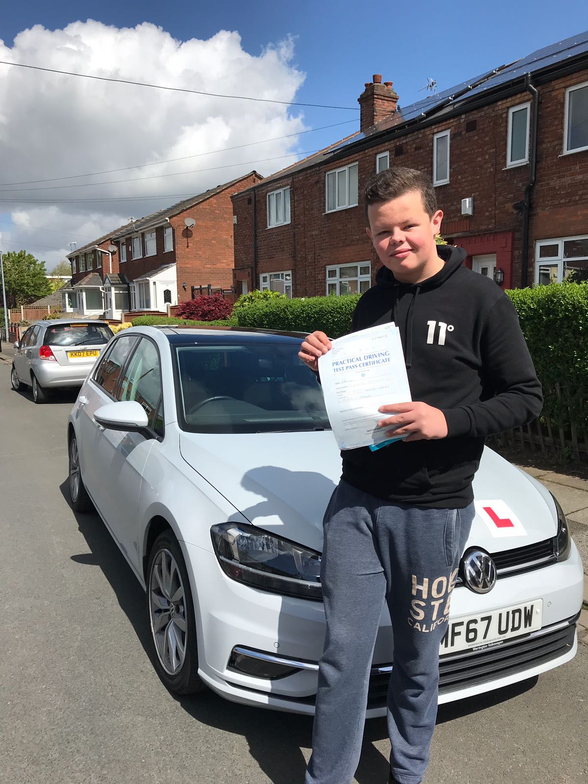 A young man is standing in front of a white car holding a certificate.
