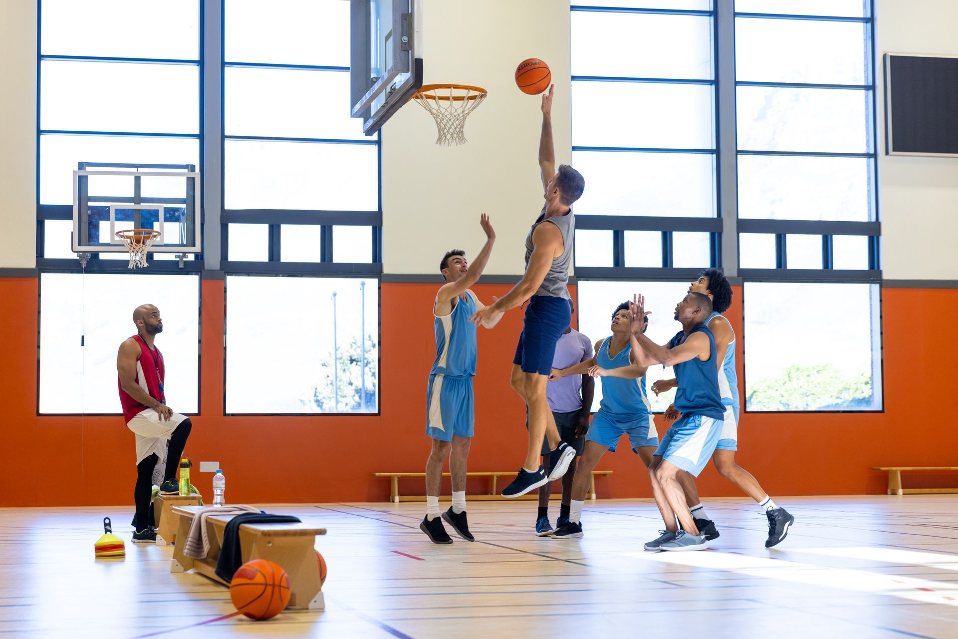 Basketball players in a gymnasium, one jumping to shoot, others defending.