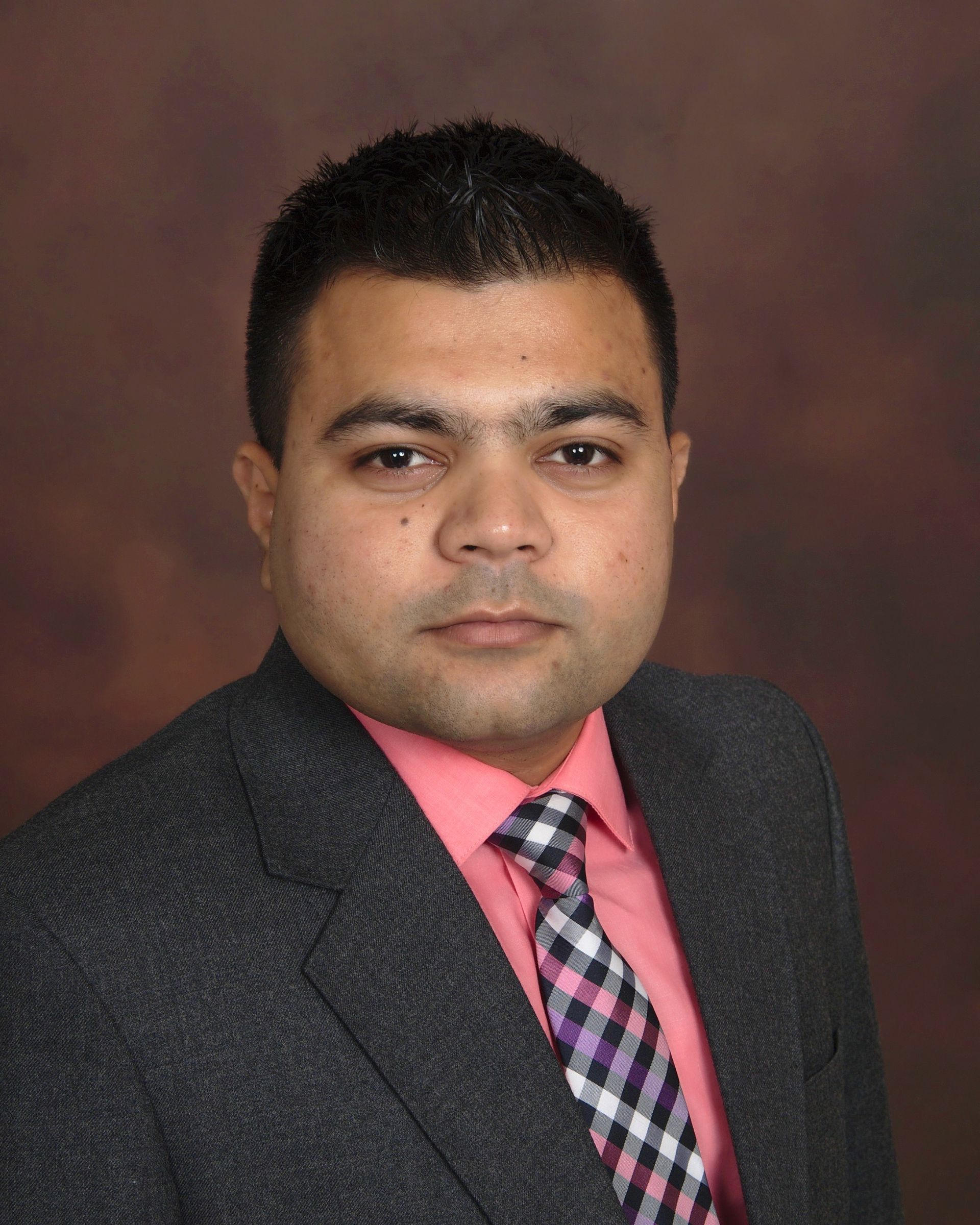 Man in a grey suit, pink shirt, and patterned tie, facing the camera with a neutral expression.