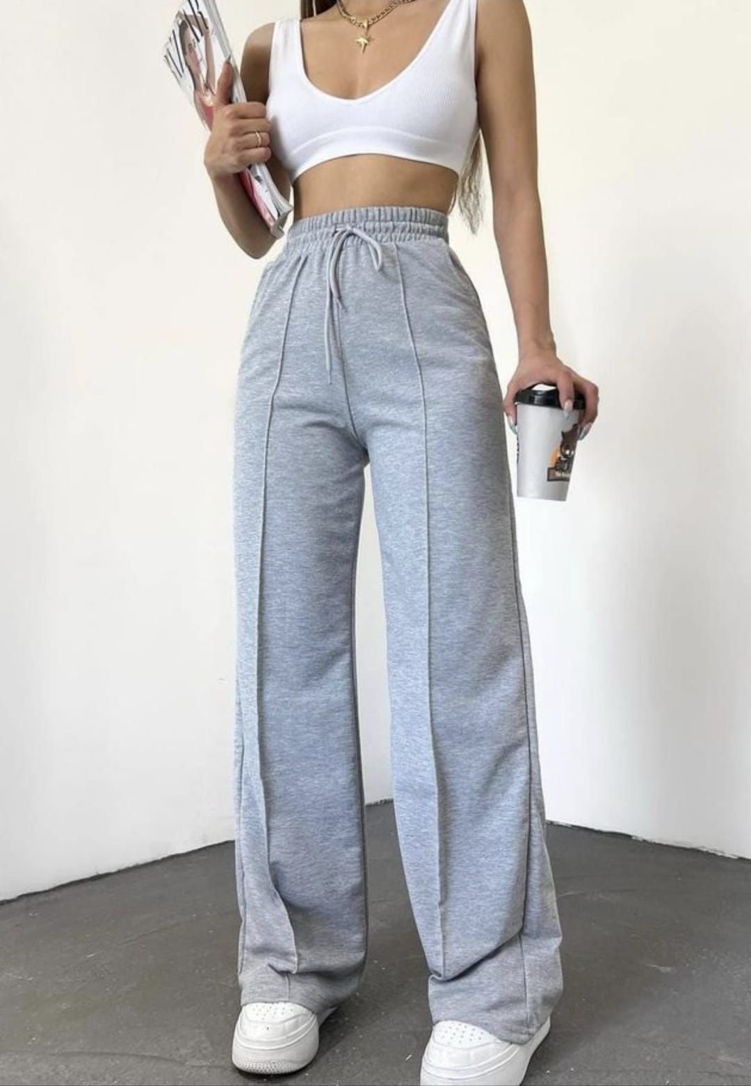 Una mujer con un top blanco y pantalones deportivos grises, sosteniendo un café y una revista, está de pie en el interior.