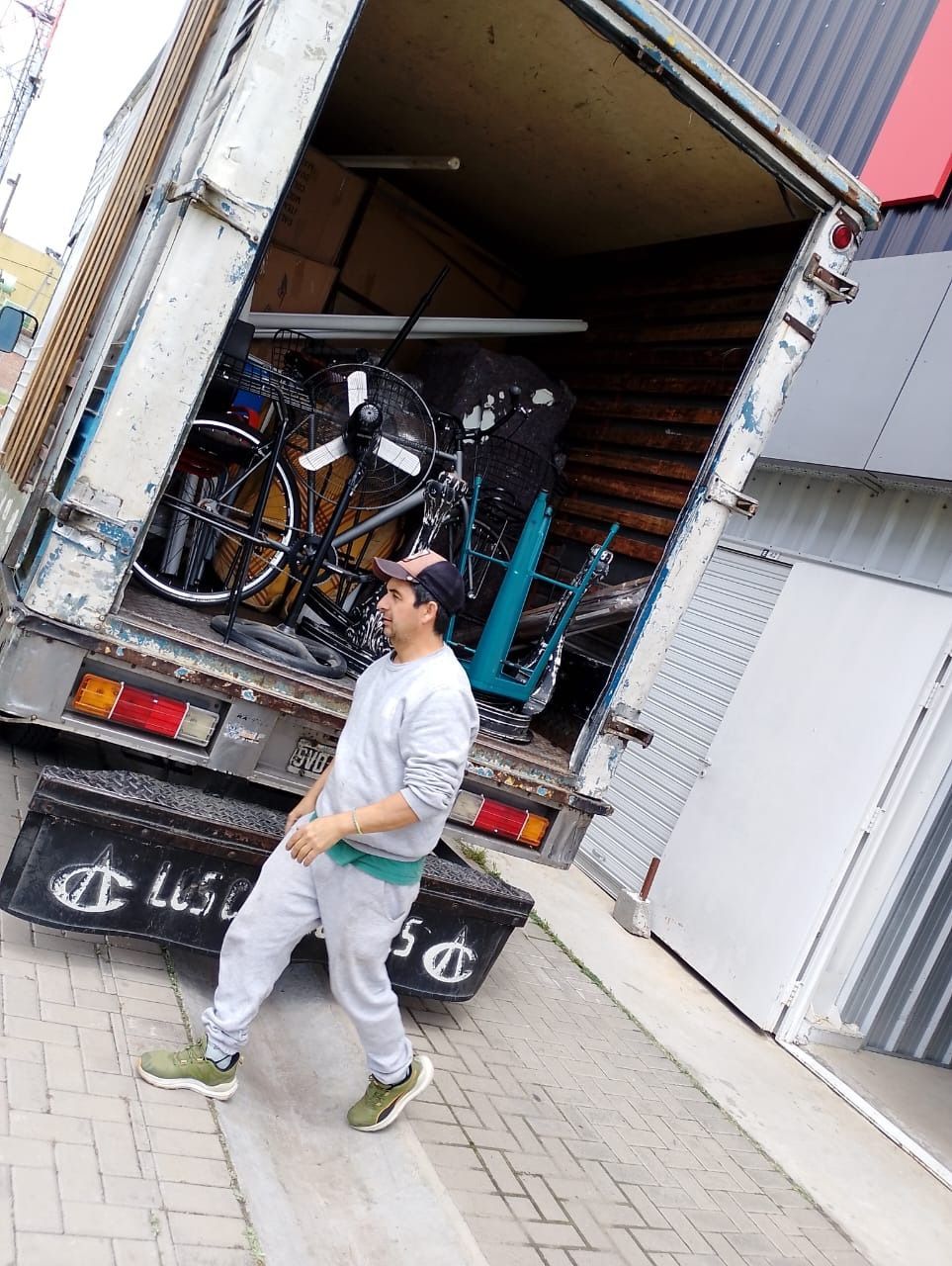 Hombre de pie junto a un camión cargado de bicicletas y otros artículos. El camión está parcialmente abierto.
