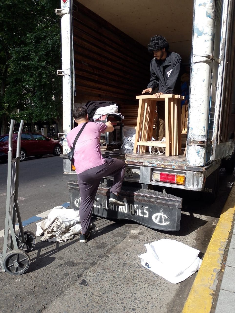 Personas cargando muebles en un camión en la calle. Una persona dentro, dos fuera, cerca de una carretilla.