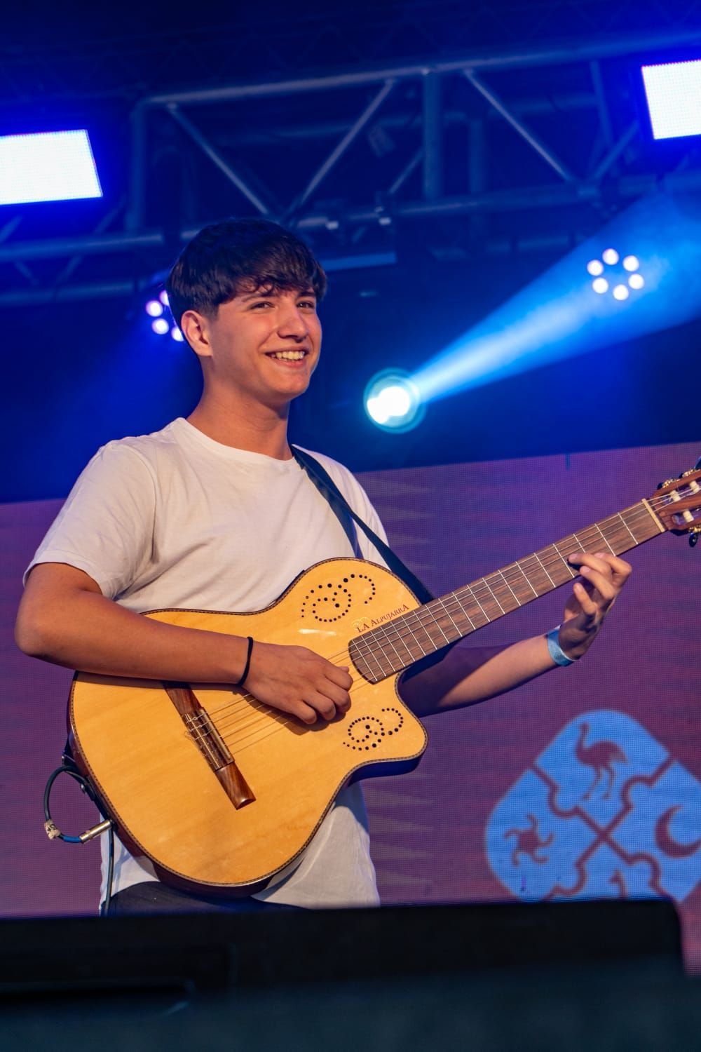 Músico sonriente tocando la guitarra acústica en el escenario, iluminación azul del escenario.