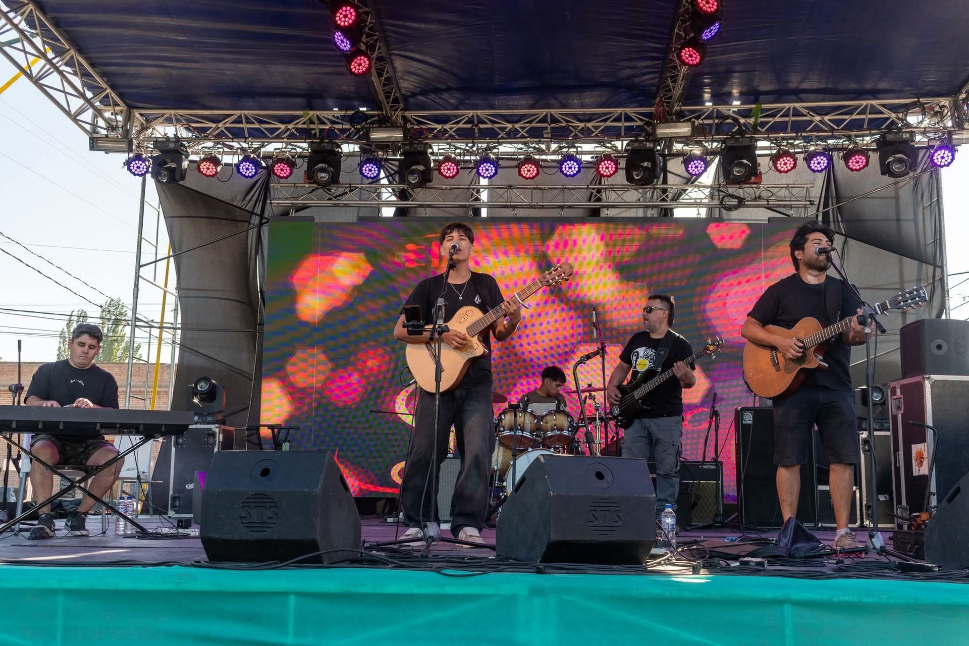 Banda actuando en un escenario al aire libre con guitarras, batería, teclado y una pantalla de vídeo.