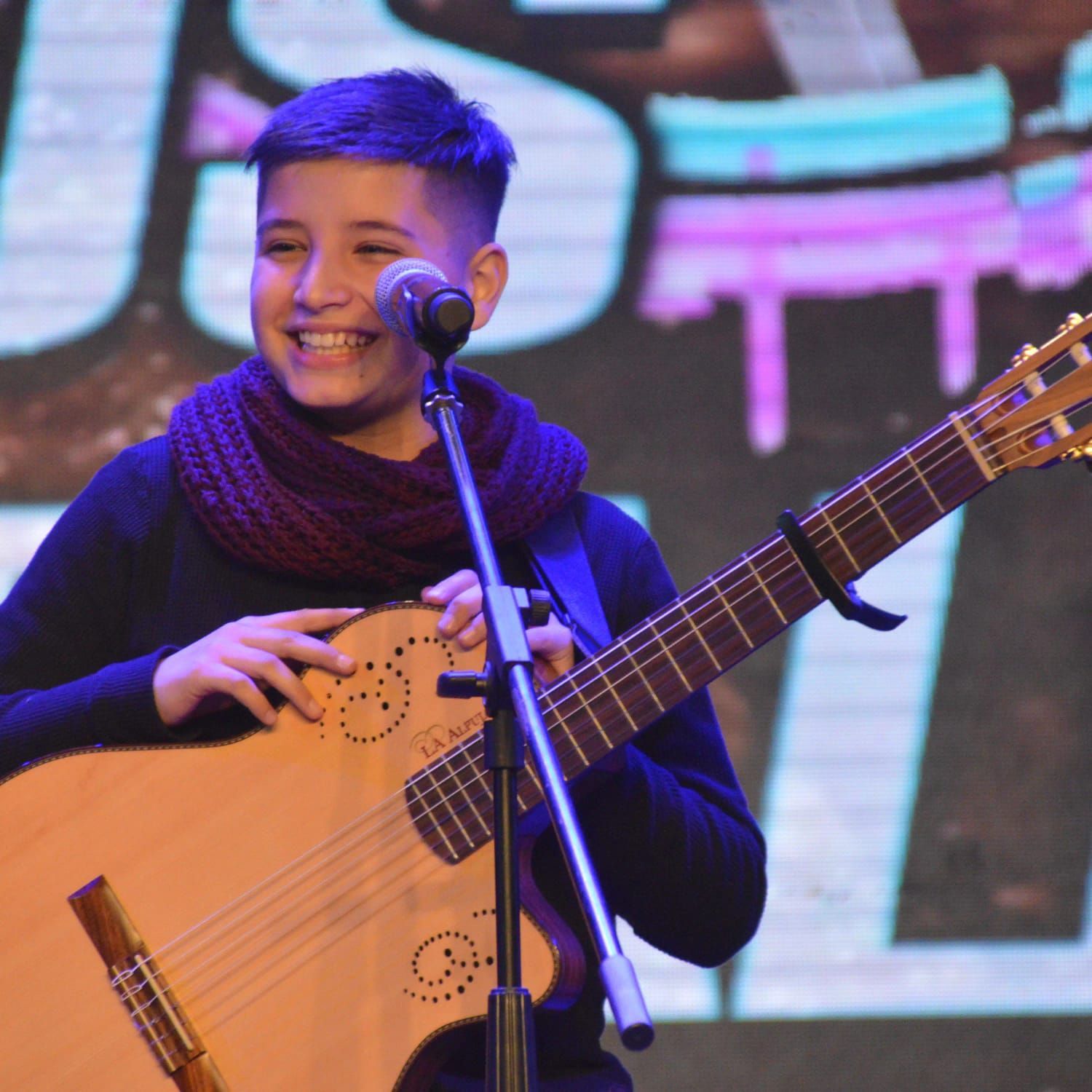 Una persona sonriente con cabello corto y azul toca la guitarra en el escenario, sosteniendo el micrófono.
