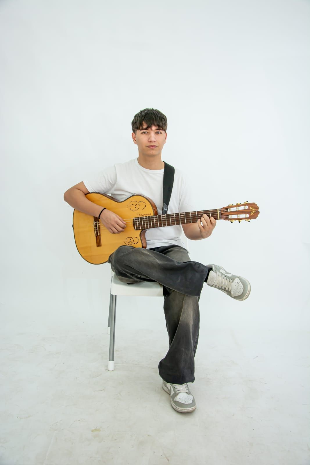 Persona joven sentada, tocando la guitarra acústica sobre un fondo blanco.