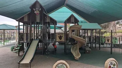 Shaded playground at Canyon Country Park with colorful play structures under a large canopy, children laughing on swings and climbing nets