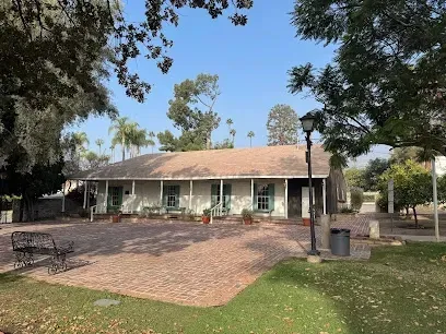 Historic white adobe house with rustic wooden beams, surrounded by lush green lawns under a bright sunny sky