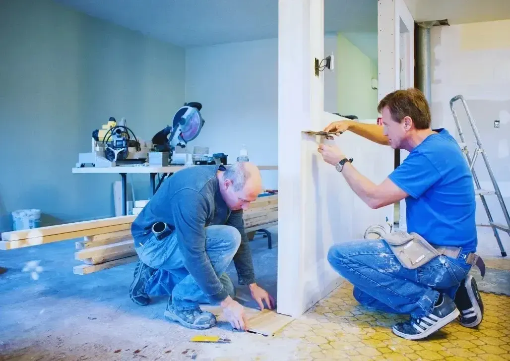 Two construction workers in blue safety suits renovating a room, with tools and exposed walls.
