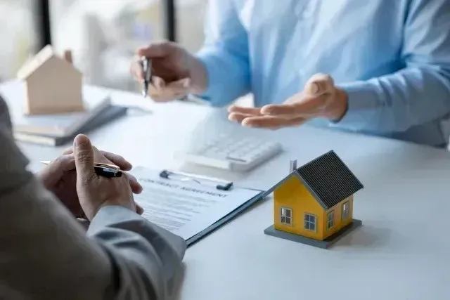 Hard Money Lender Los Angeles - Two people discussing over a house model and contract on a table. One person gestures while the other holds a pen near the contract.
