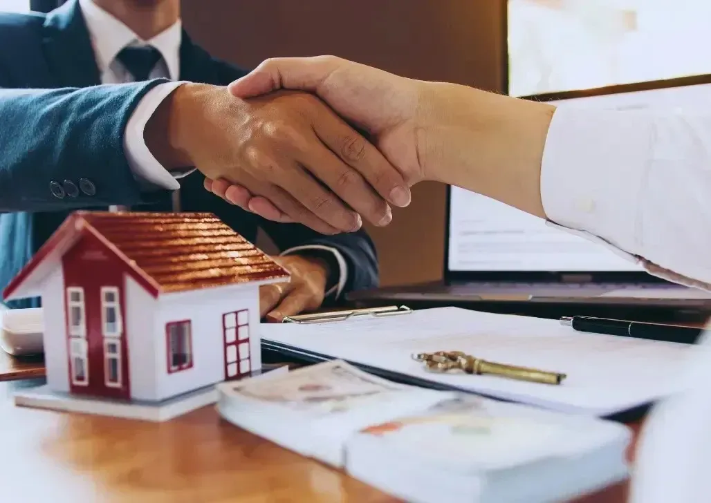 Real estate agent and client shaking hands at table with contract papers and miniature house model