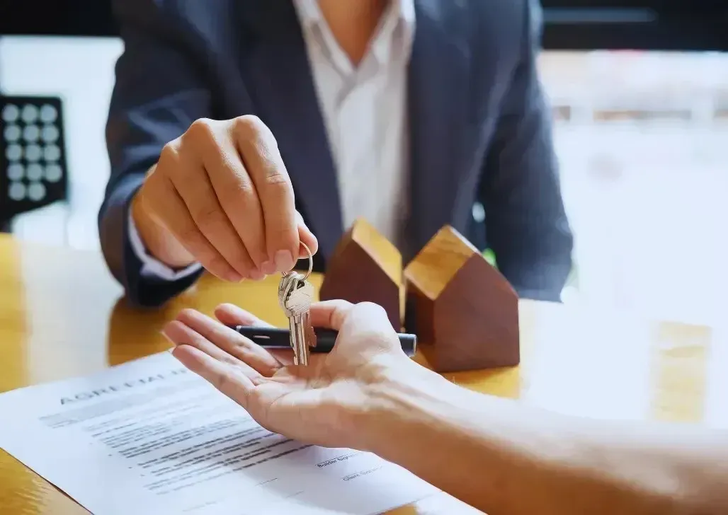 Real estate agent handing house keys to smiling client, with contract documents and miniature home model on table