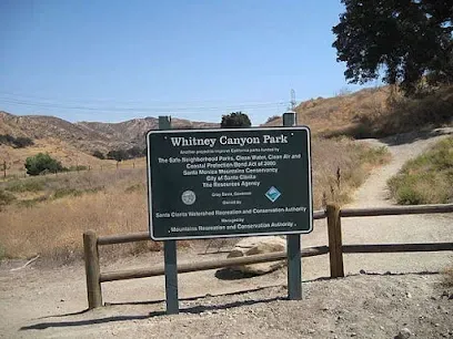 Sun-dappled hiking trail at Whitney Canyon Park, with a wooden information kiosk and rustic split-rail fences lining the dusty path