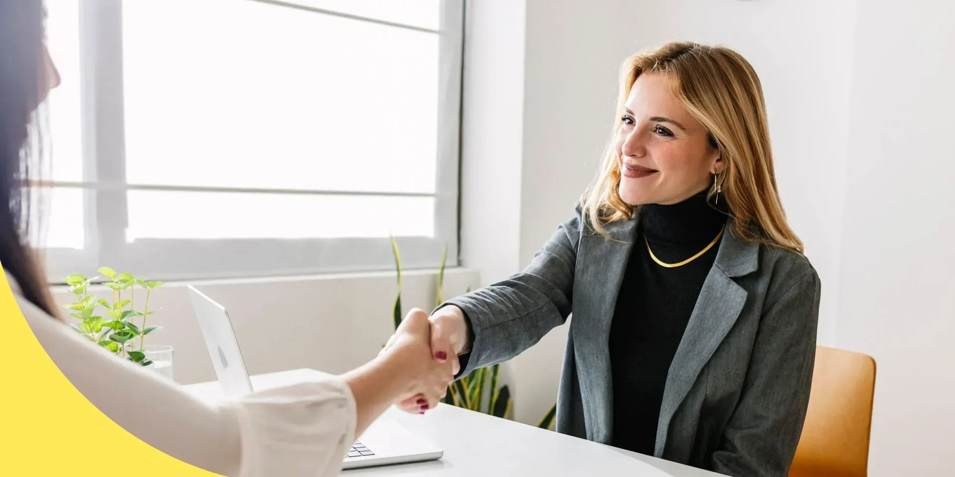 Smiling professional woman shaking hands with a client across a desk, open laptop and documents visible during their discussion