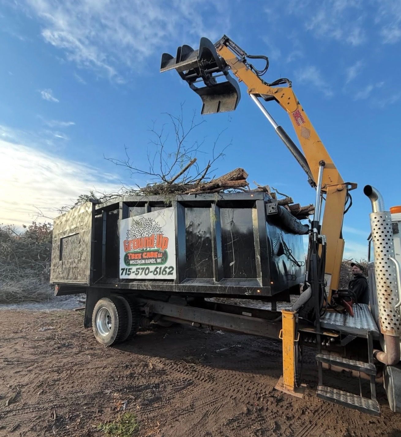 A skid steer with a tree shear attachment cutting tree branches. An orange-vested person stands nearby.