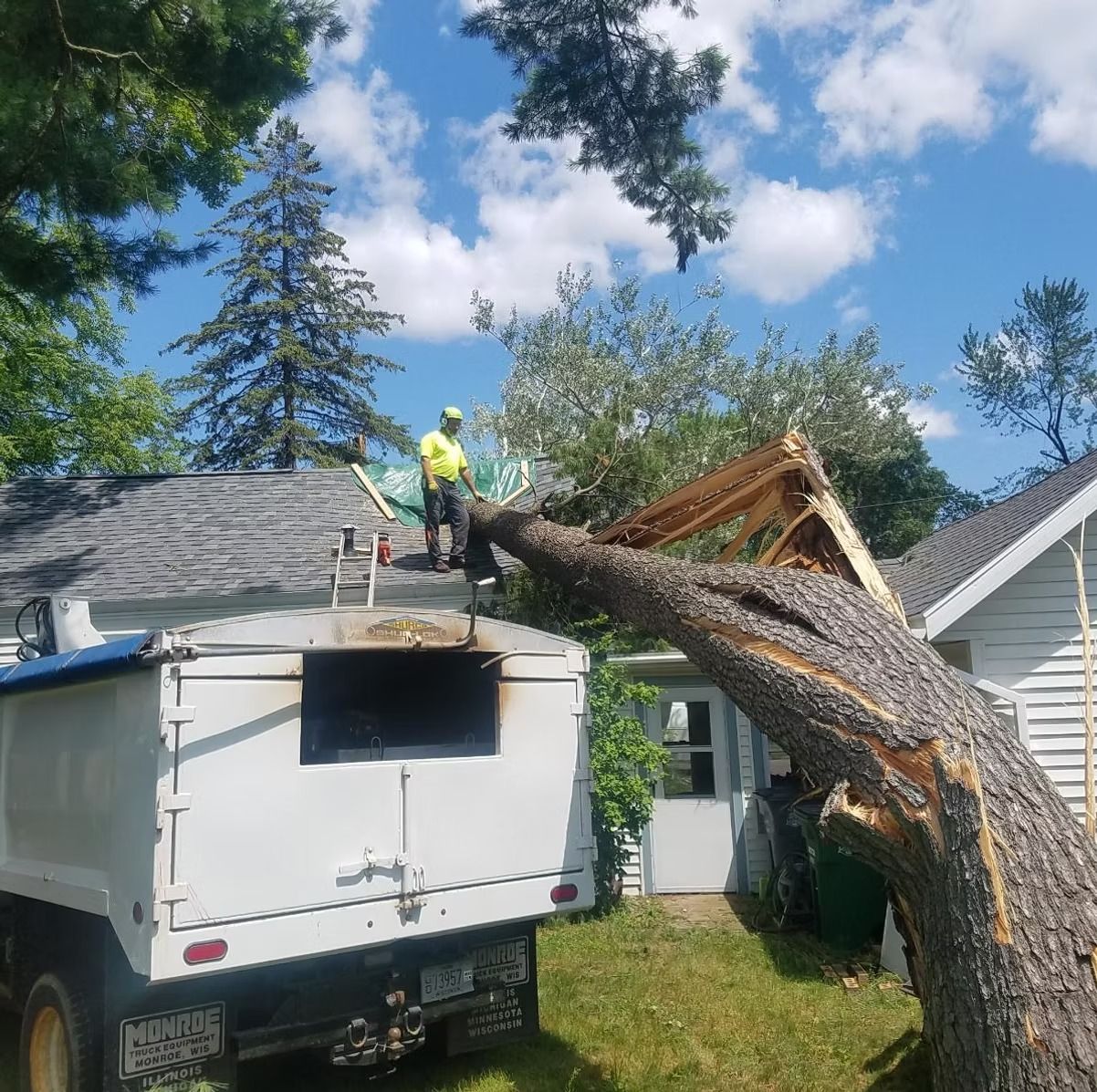 Tree removal crew on roof, cutting fallen tree from house. Truck, blue sky.