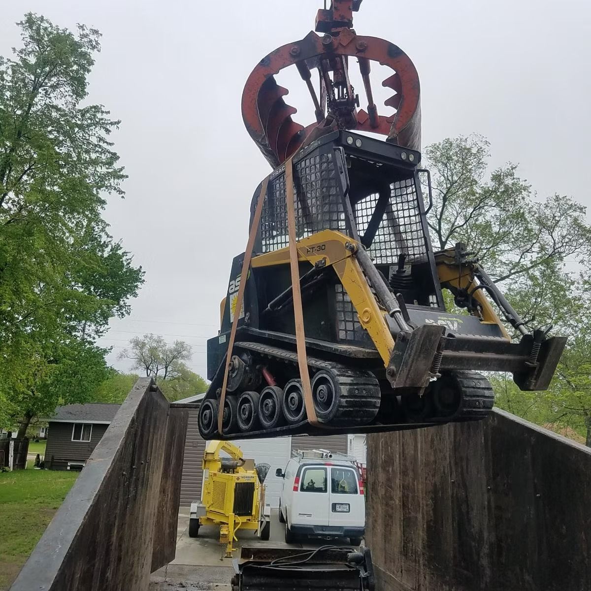 A skid steer is lifted into a dump truck by an overhead claw. A yellow chipper and white van are visible.