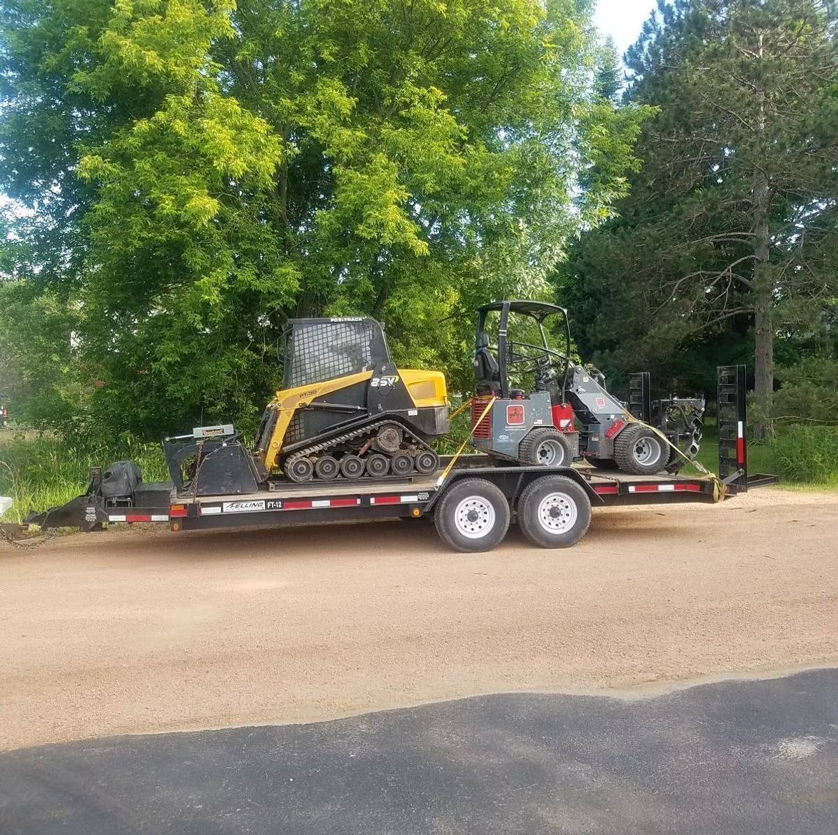 A trailer loaded with two mini excavators, one yellow, the other gray, parked on gravel. Green trees in the background.
