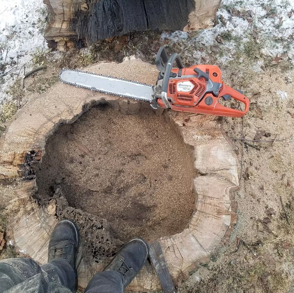 Chainsaw on a tree stump, surrounded by sawdust. A person's feet in boots are visible. Snowy ground.
