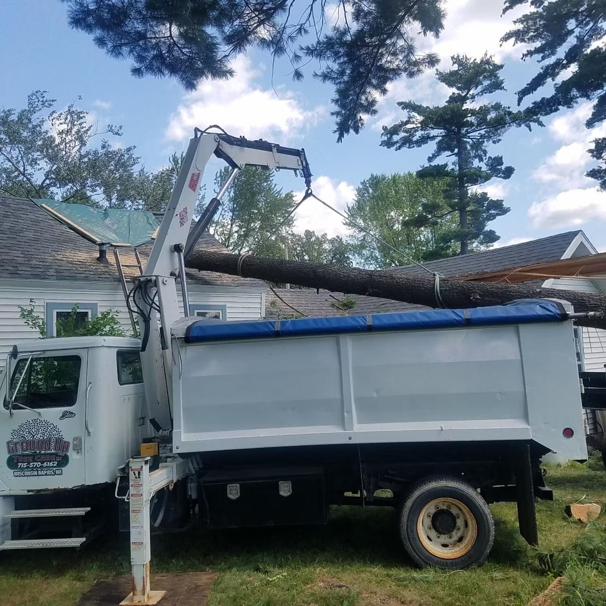 White tree service truck with crane loading a large log; next to a house.