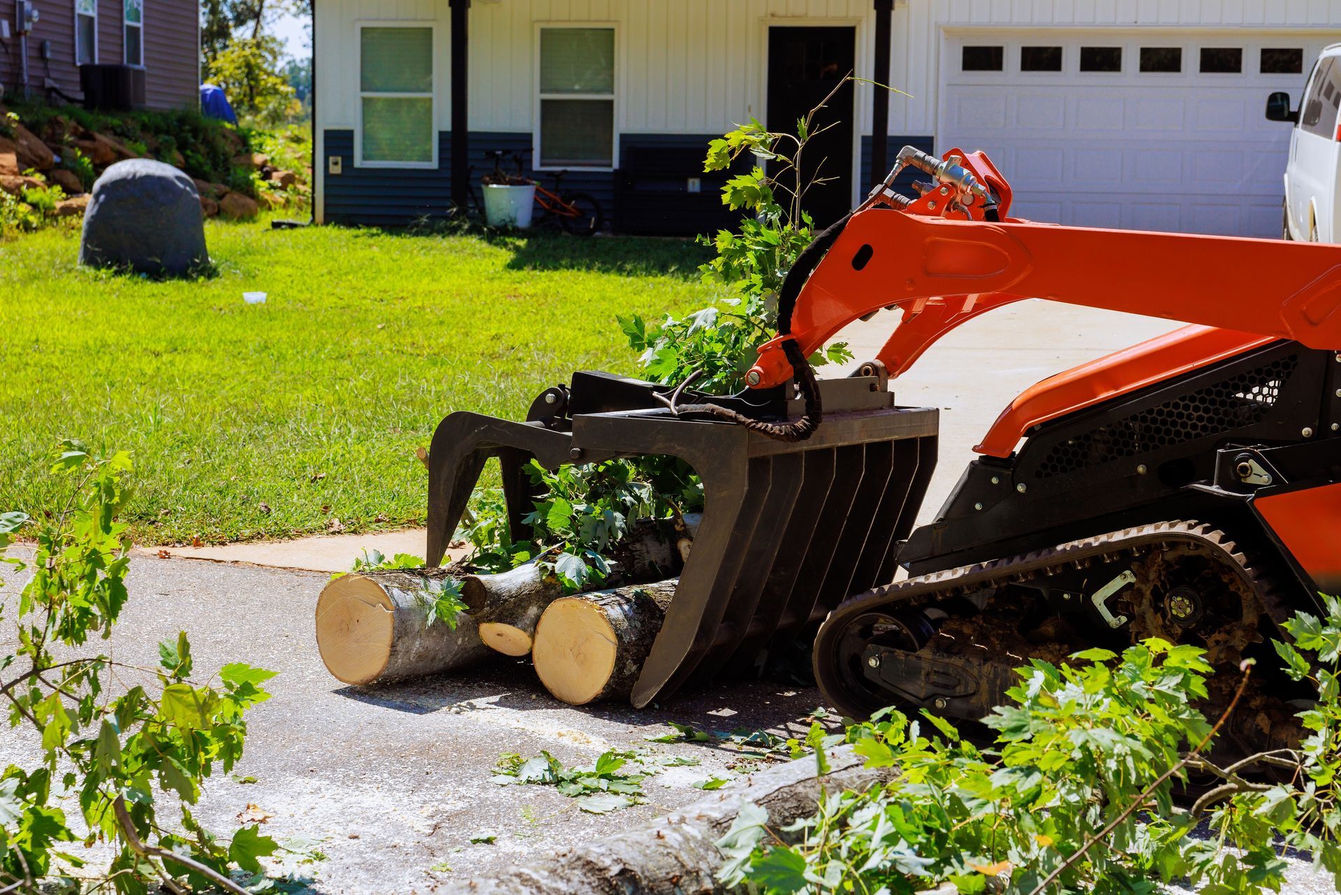 Orange mini skid steer with grapple arms lifting logs on a driveway, near a house with green grass.