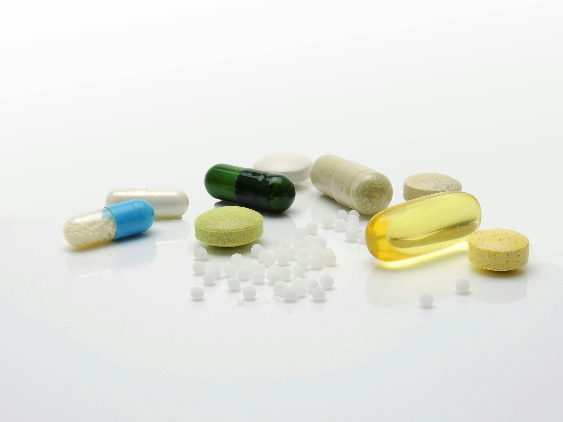 A scattering of various colorful pharmaceutical pills, capsules, and small white granules on a plain white surface.