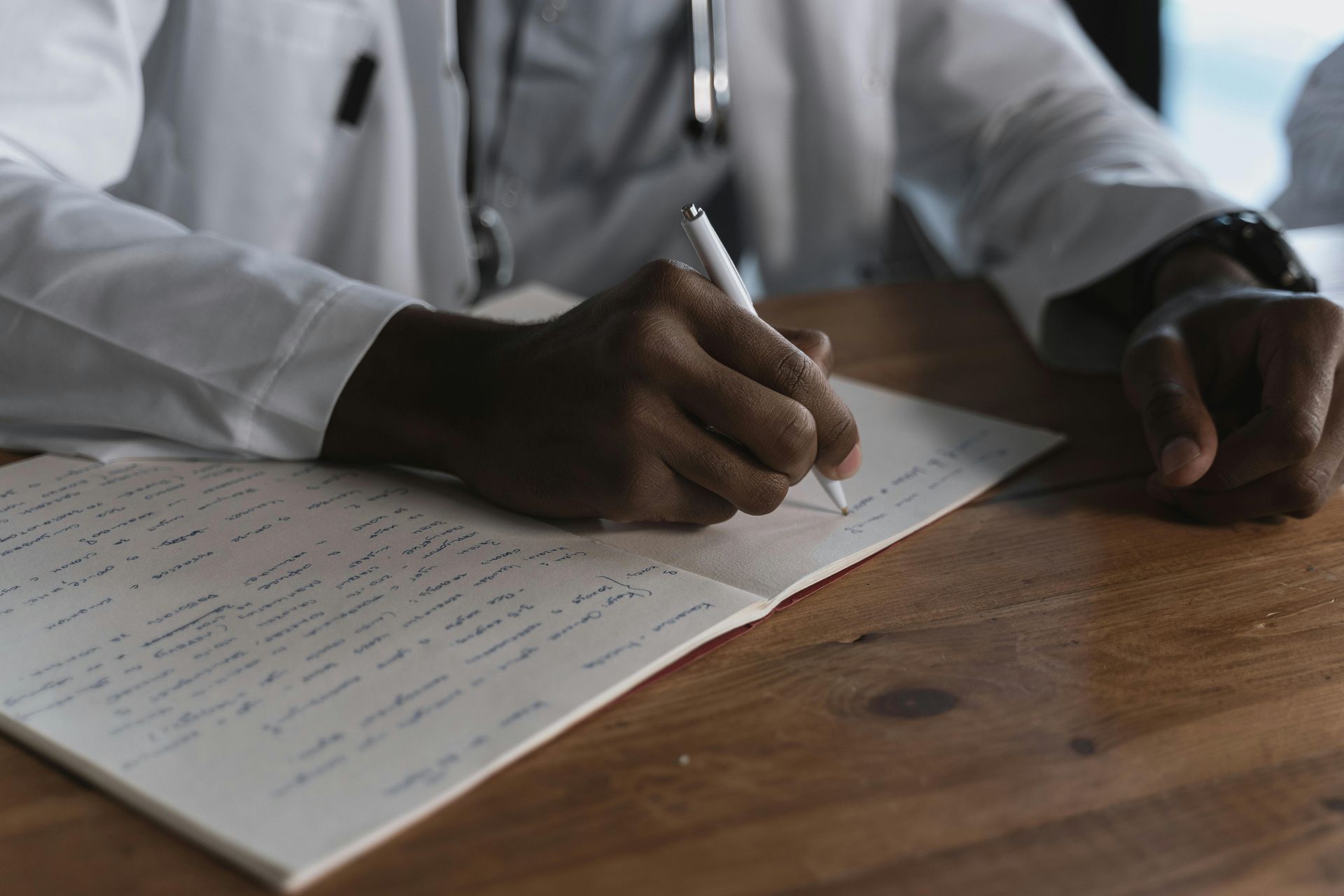 A person in a white medical coat writes in a notebook at a wooden table.