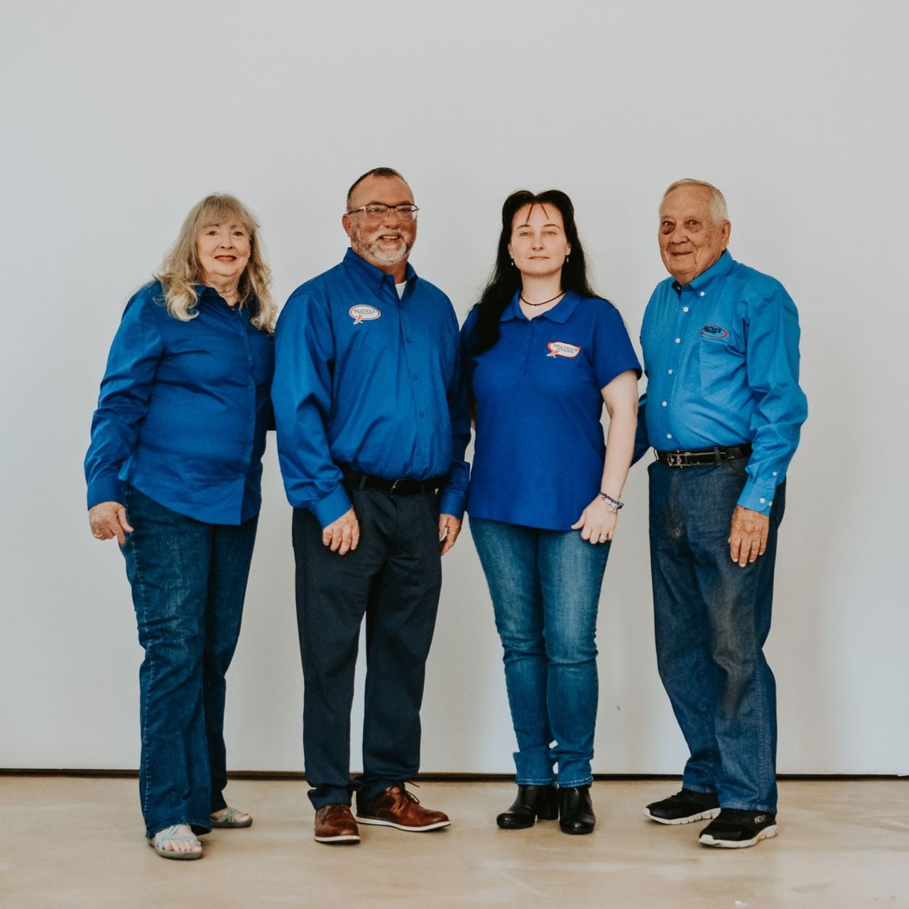 Four people standing in a row against a plain white wall, all wearing blue shirts and jeans.