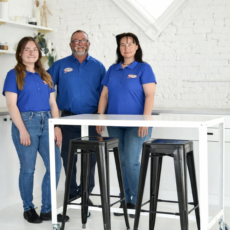 Three people in matching blue polo shirts stand behind a white table with two black stools in a bright, modern studio.