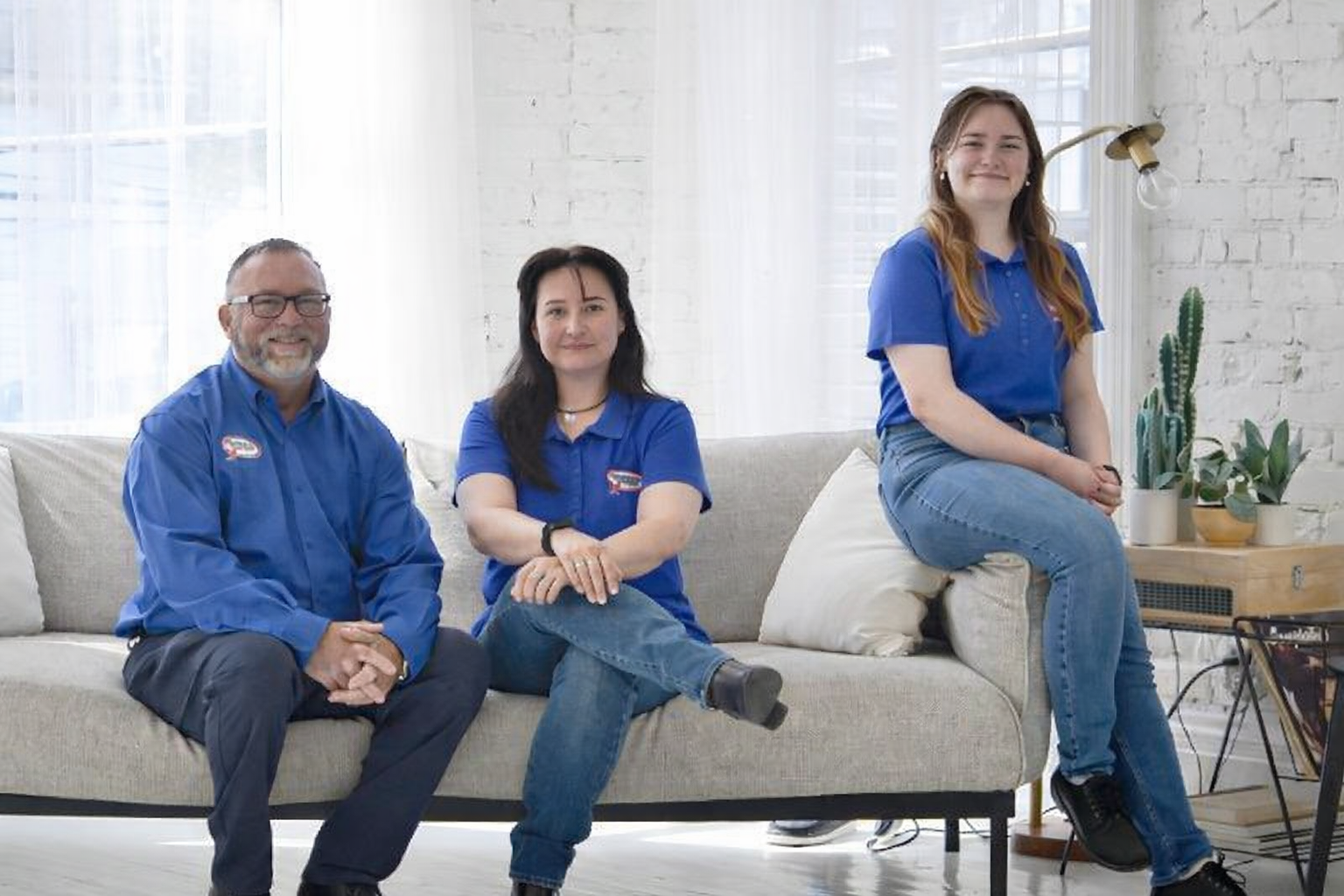 Three people in matching blue shirts sit on a neutral couch in a sunlit room with brick walls.