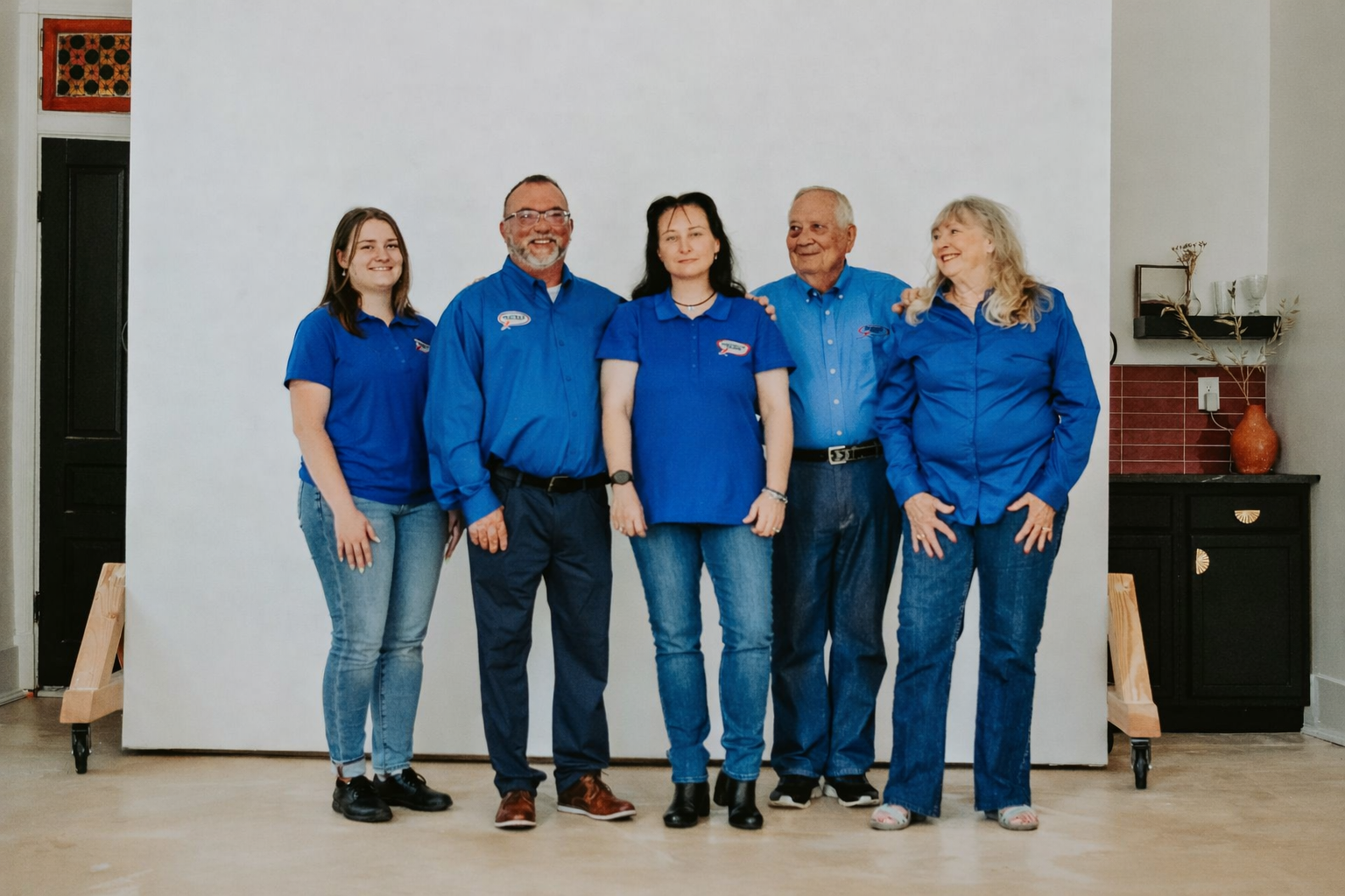 Five people standing in a row against a white backdrop, all wearing matching royal blue shirts and jeans.