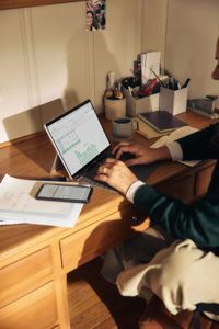 A person sits at a wooden desk working on a tablet displaying a chart, with stationery and a smartphone nearby.